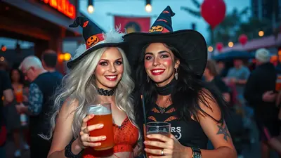 Two women in witch costumes, wearing pointed hats and holding drinks, smile at the camera at an outdoor Halloween event.