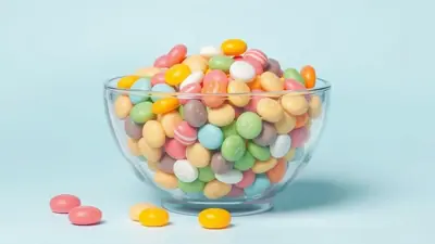 A clear glass bowl overflowing with colorful, pastel-colored candies on a light blue background. A few candies are scattered on the surface in front of the bowl.
