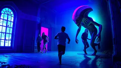 A group of people are running away from a tall, menacing alien creature in a dimly lit room with blue and purple lighting. The alien has a distinctive head shape and appears to be chasing the people.