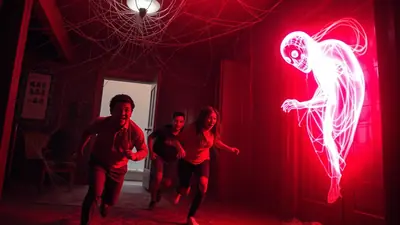 Three people are running away from a glowing, skeletal apparition in a room decorated with fake spiderwebs and bathed in red light.