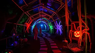 A dark, spooky tunnel decorated for Halloween with neon spider and pumpkin decorations, and two people walking through it.