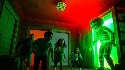 A group of people are running away from a glowing green alien in a hallway with red and green lighting.