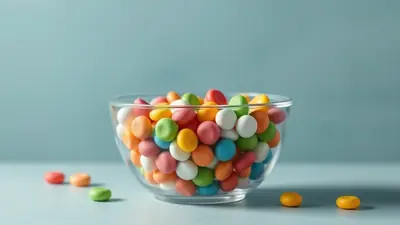 A clear glass bowl filled with colorful, round candies sits on a light blue surface against a soft blue background. A few candies are scattered on the surface in front of the bowl.
