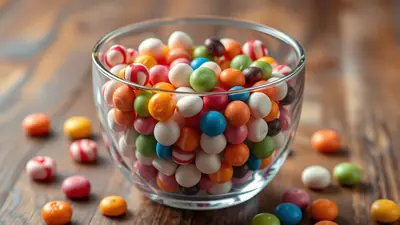 A clear glass bowl filled with colorful round candies, including white, orange, green, blue, pink, and red striped candies, sits on a wooden surface. Several candies are scattered around the bowl on the surface.