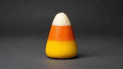 A single piece of candy corn with distinct yellow, orange, and white stripes sits on a dark gray surface against a dark gray background.